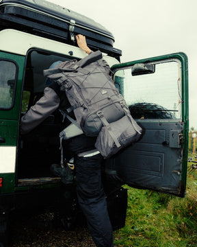 Person klettert mit großem grauem Johnny Urban Allen XL Travel Reiserucksack in grünes Geländefahrzeug