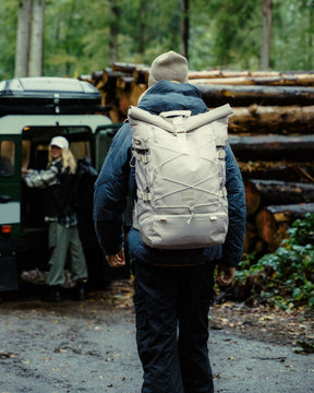 Mann trägt beigen Johnny Urban Reiserucksack 'Allen XL Travel' im Wald, Frau steht neben grünem Geländewagen