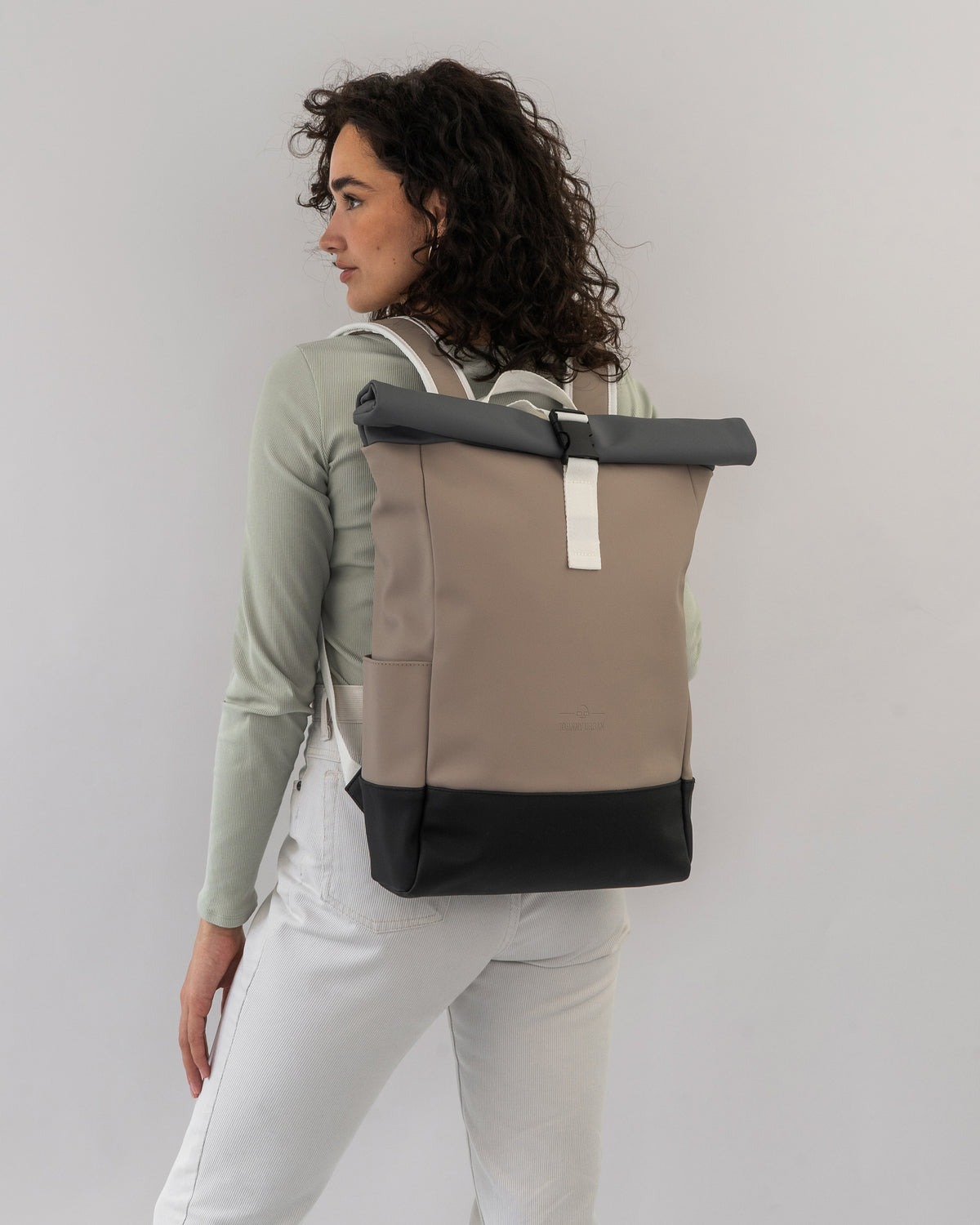 Frau mit Rolltop-Rucksack 'Harvey Medium' in Beige, Grau und Schwarz, trägt ihn an den weißen Hosen und grünem Oberteil rückseitig