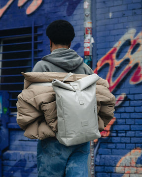 Mann trägt beigen Rolltop Rucksack 'Robin Medium' von Johnny Urban und beige Jacke vor blauem Graffiti-Hintergrund