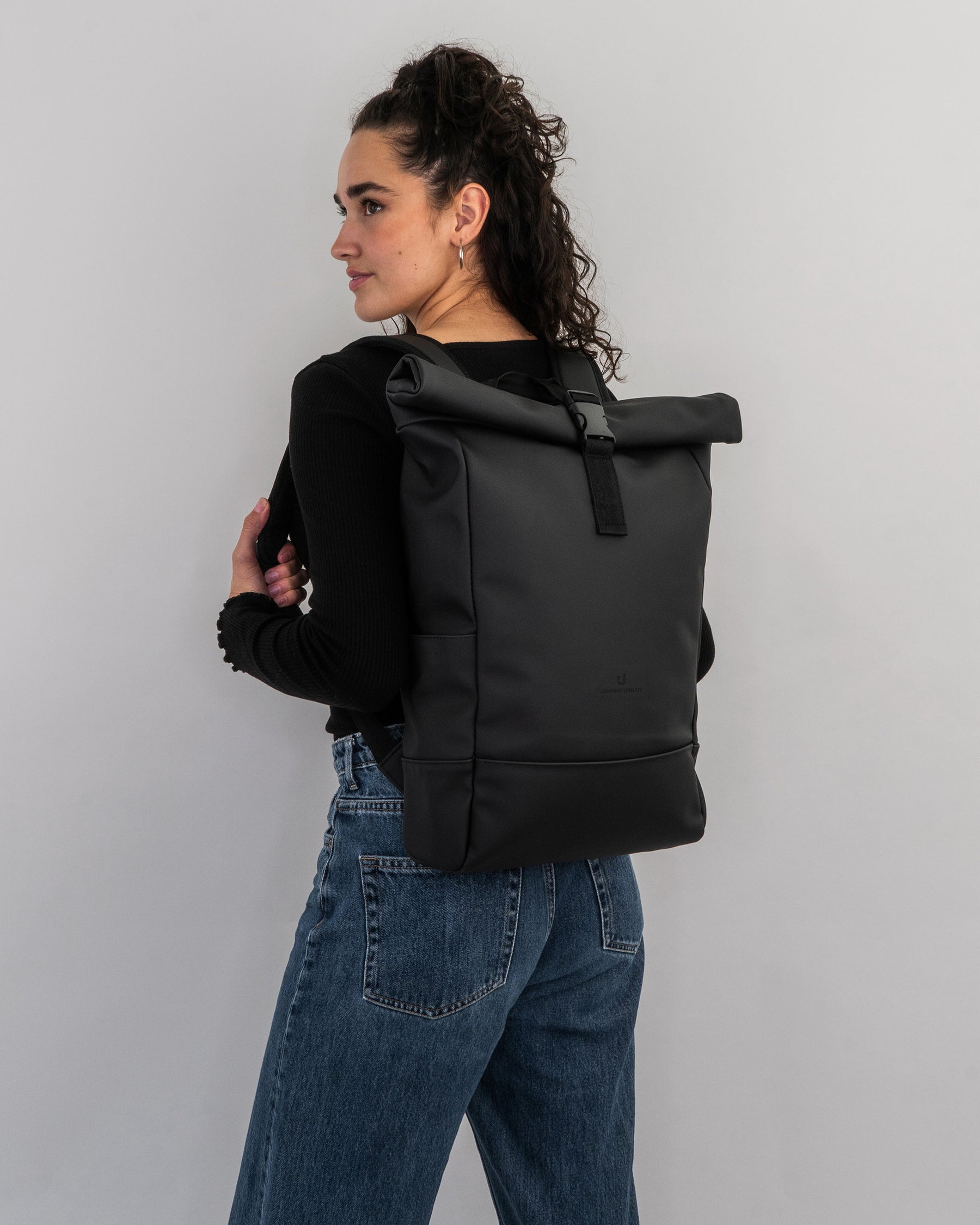 Frau mit lockigem Haar trägt schwarzen Rolltop-Rucksack 'Harvey Medium' von Johnny Urban an schwarzem Oberteil und blauen Jeans