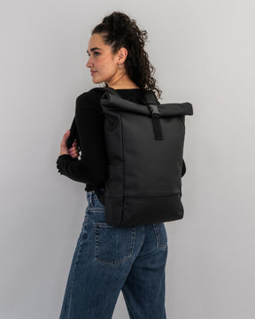 Frau mit lockigem Haar trägt schwarzen Rolltop-Rucksack 'Harvey Medium' von Johnny Urban an schwarzem Oberteil und blauen Jeans
