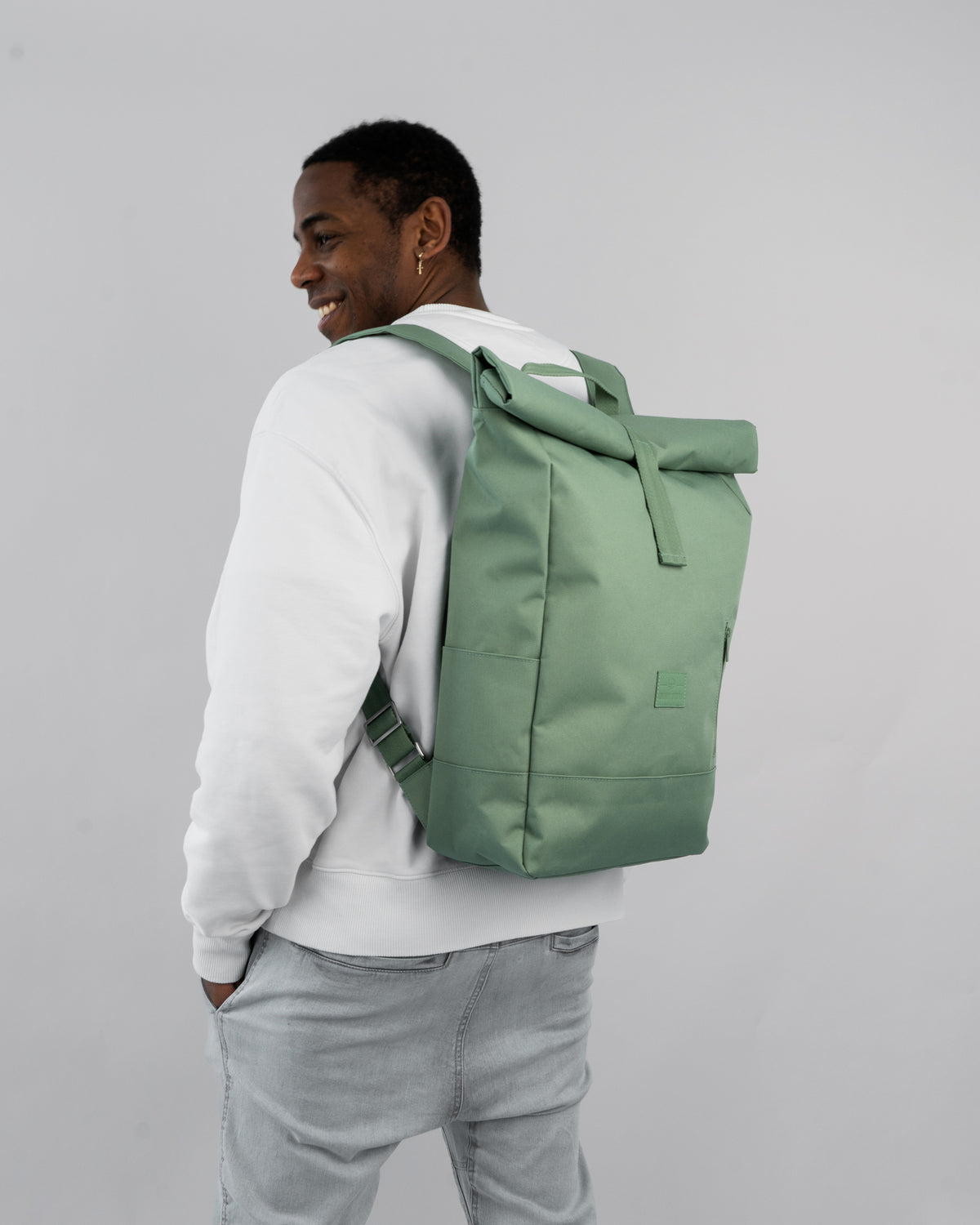 Mann trägt grünen Rolltop-Rucksack 'Robin Large' von Johnny Urban, kombiniert mit weißem Pullover und hellen Jeans