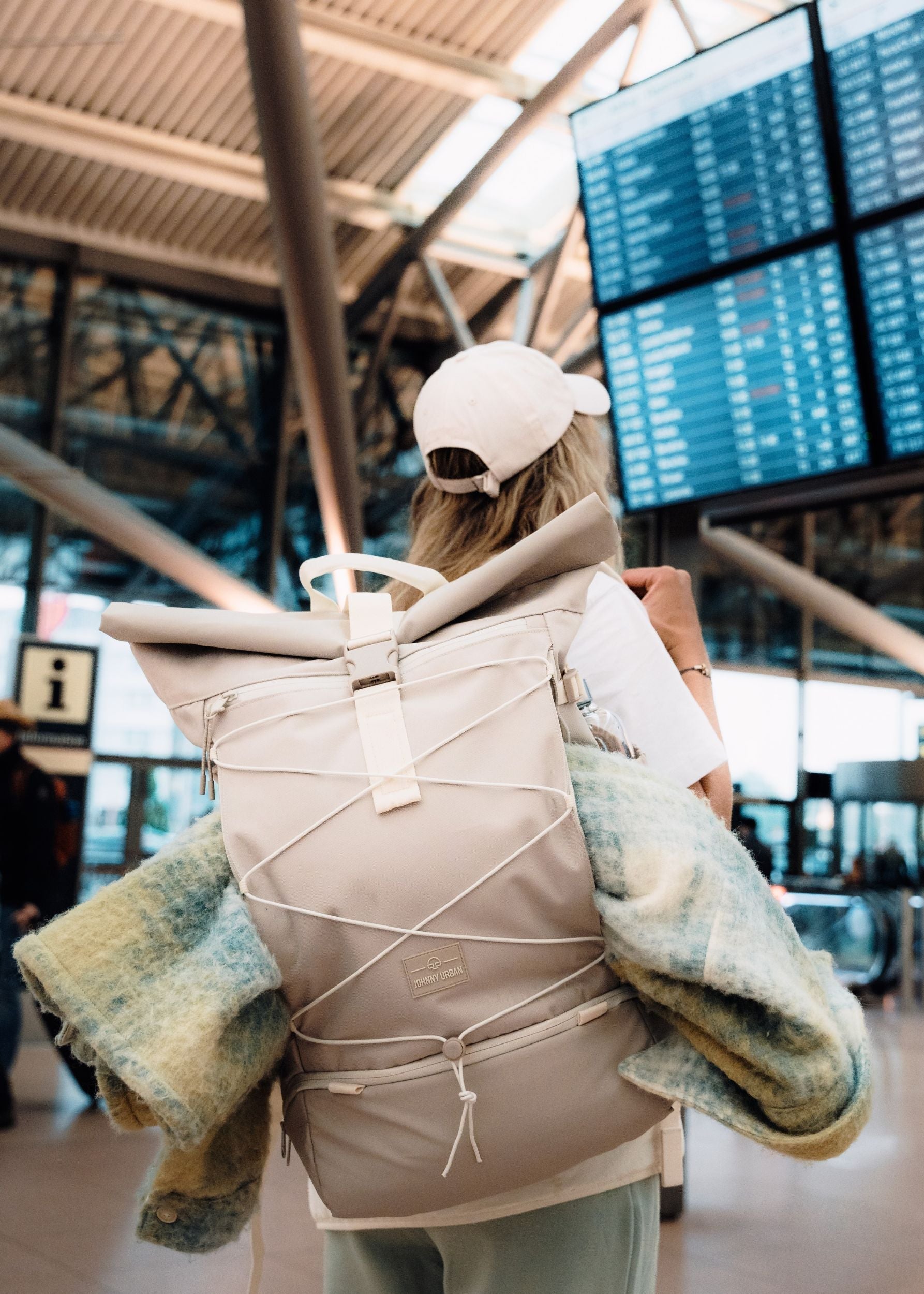 Frau an einem Flughafen mit beigefarbenem Rolltop-Reiserucksack ‚Allen Large Travel‘ von Johnny Urban auf dem Rücken