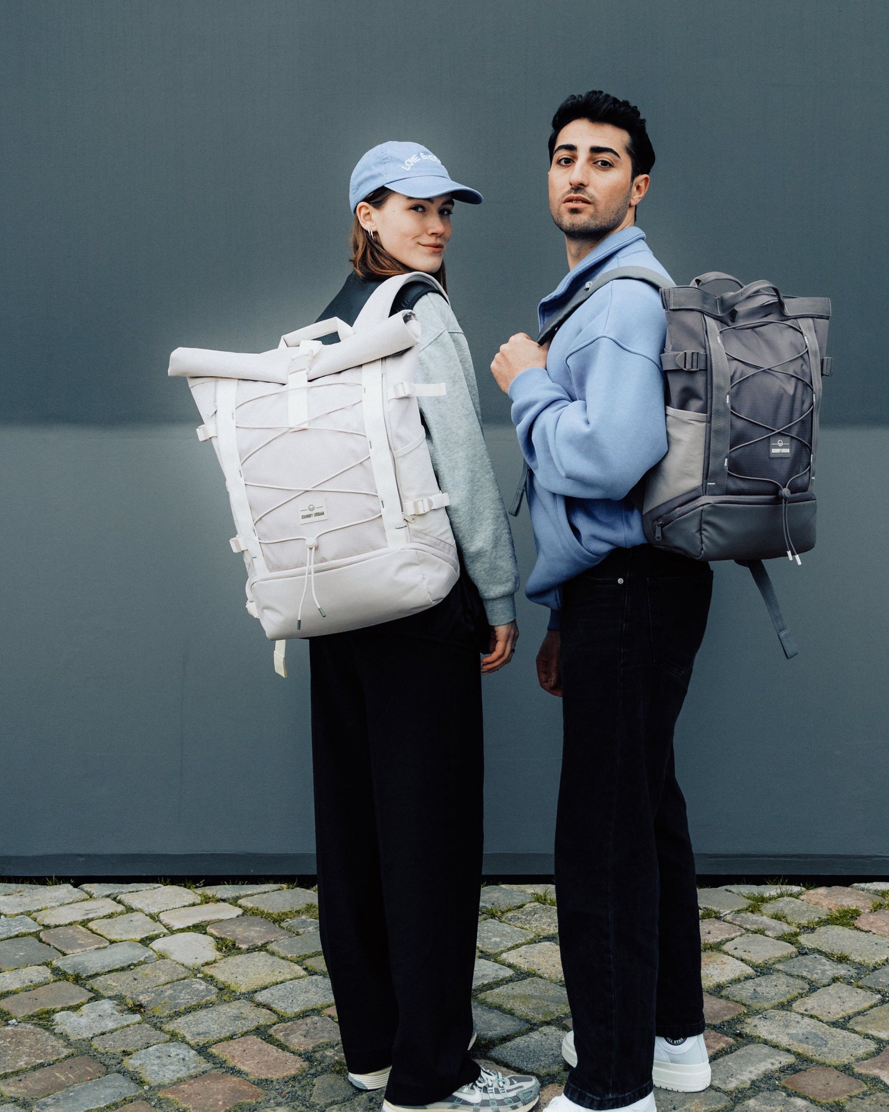 Zwei Personen tragen Rolltop Rucksäcke 'Allen Large Move', eine beige und eine graue, auf gepflastertem Boden vor grauer Wand
