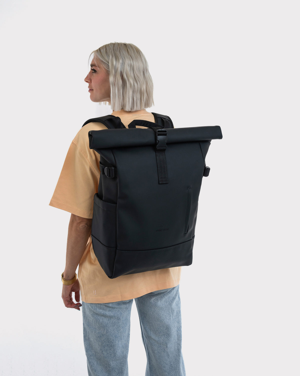 Frau mit kurzem blondem Haar trägt schwarzen Rolltop Rucksack 'Harvey Large' von hinten in lässiger Kleidung