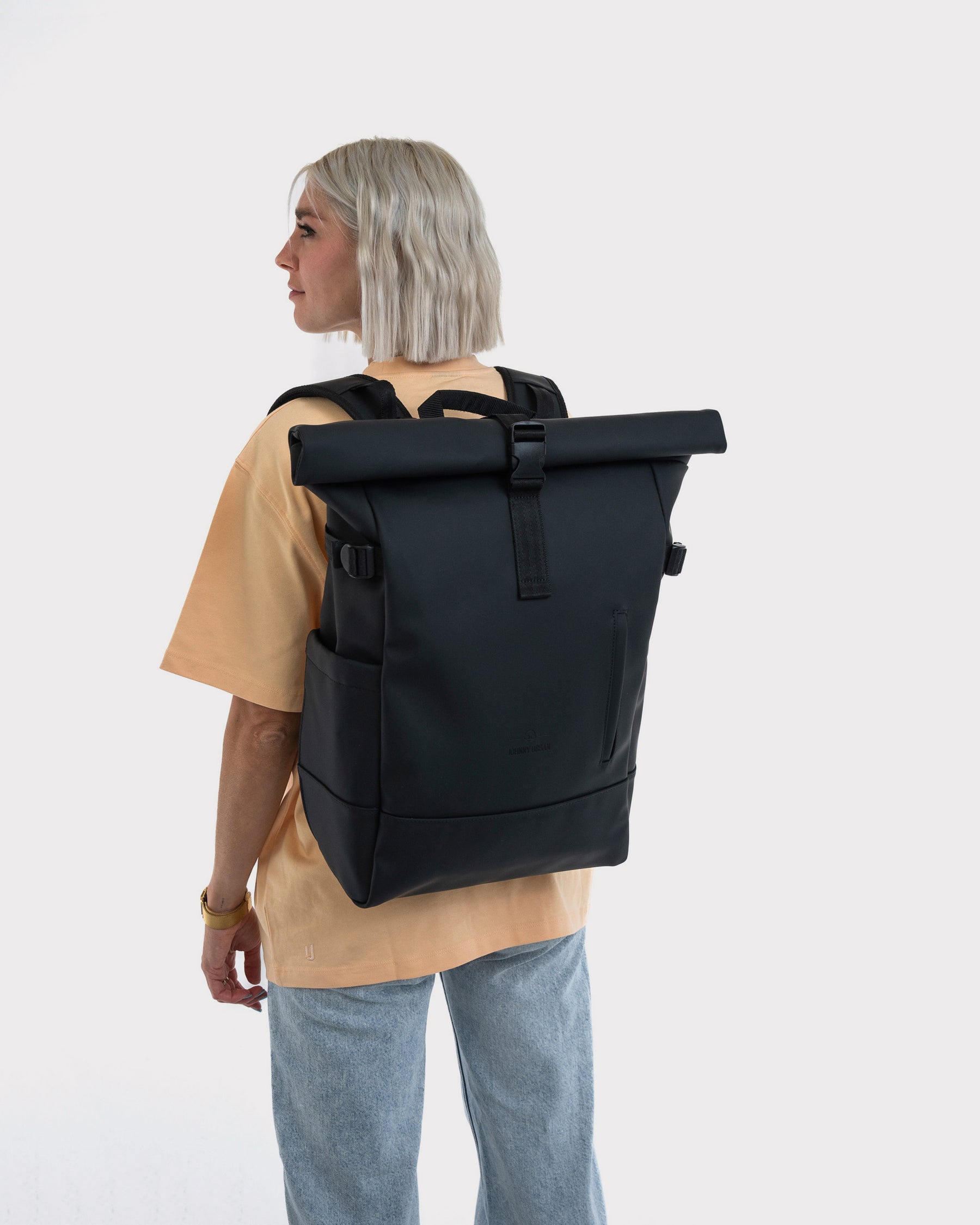 Frau mit kurzem blondem Haar trägt schwarzen Rolltop Rucksack 'Harvey Large' von hinten in lässiger Kleidung