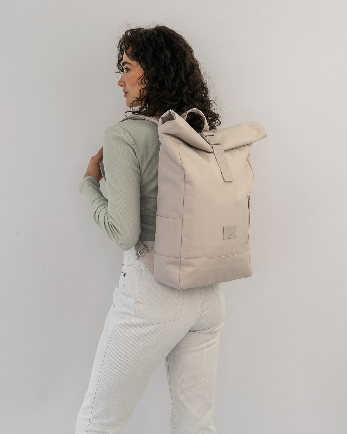 Frau mit lockigen Haaren trägt hellbeigen Rolltop Rucksack 'Robin Medium' vor weißem Hintergrund
