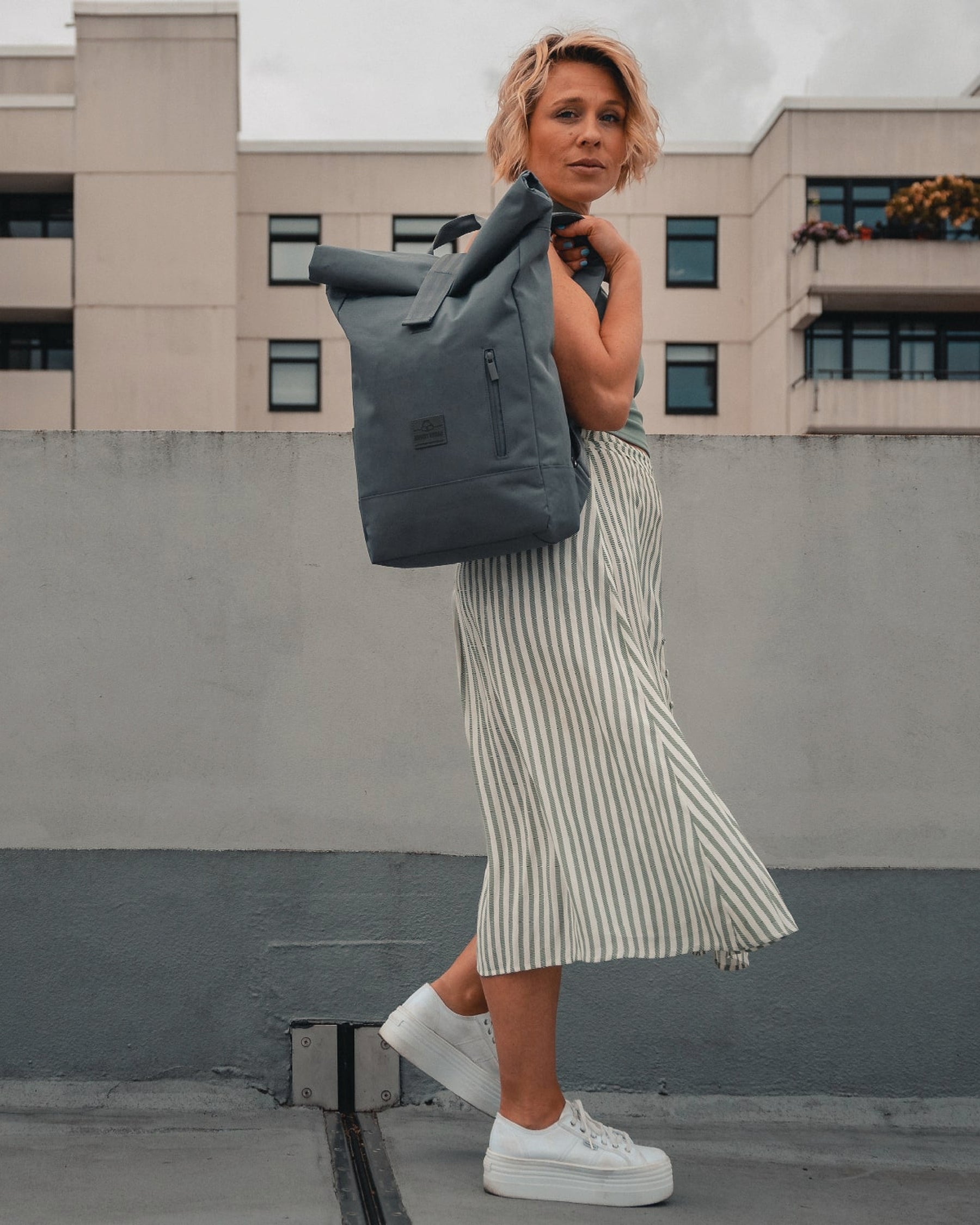 Frau trägt dunkelgrauen Rolltop Rucksack ‚Robin Medium‘, gestreifter Midirock, weiße Sneaker in urbaner Umgebung