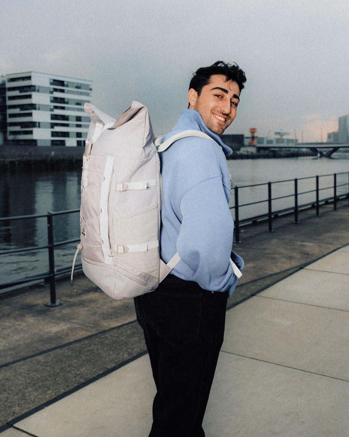 Mann trägt beigen Rolltop Rucksack 'Allen Large Move' und hellblauen Pullover am Wasser in urbaner Umgebung