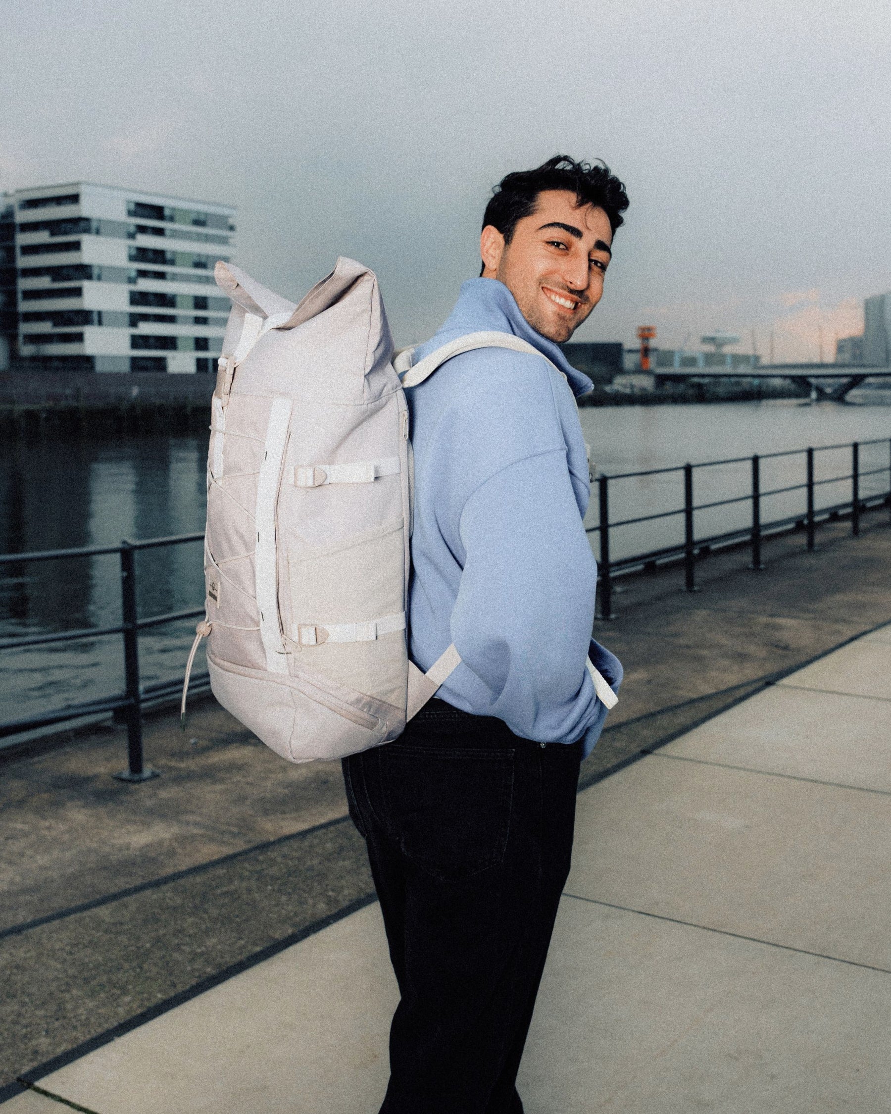 Mann trägt beigen Rolltop Rucksack 'Allen Large Move' und hellblauen Pullover am Wasser in urbaner Umgebung
