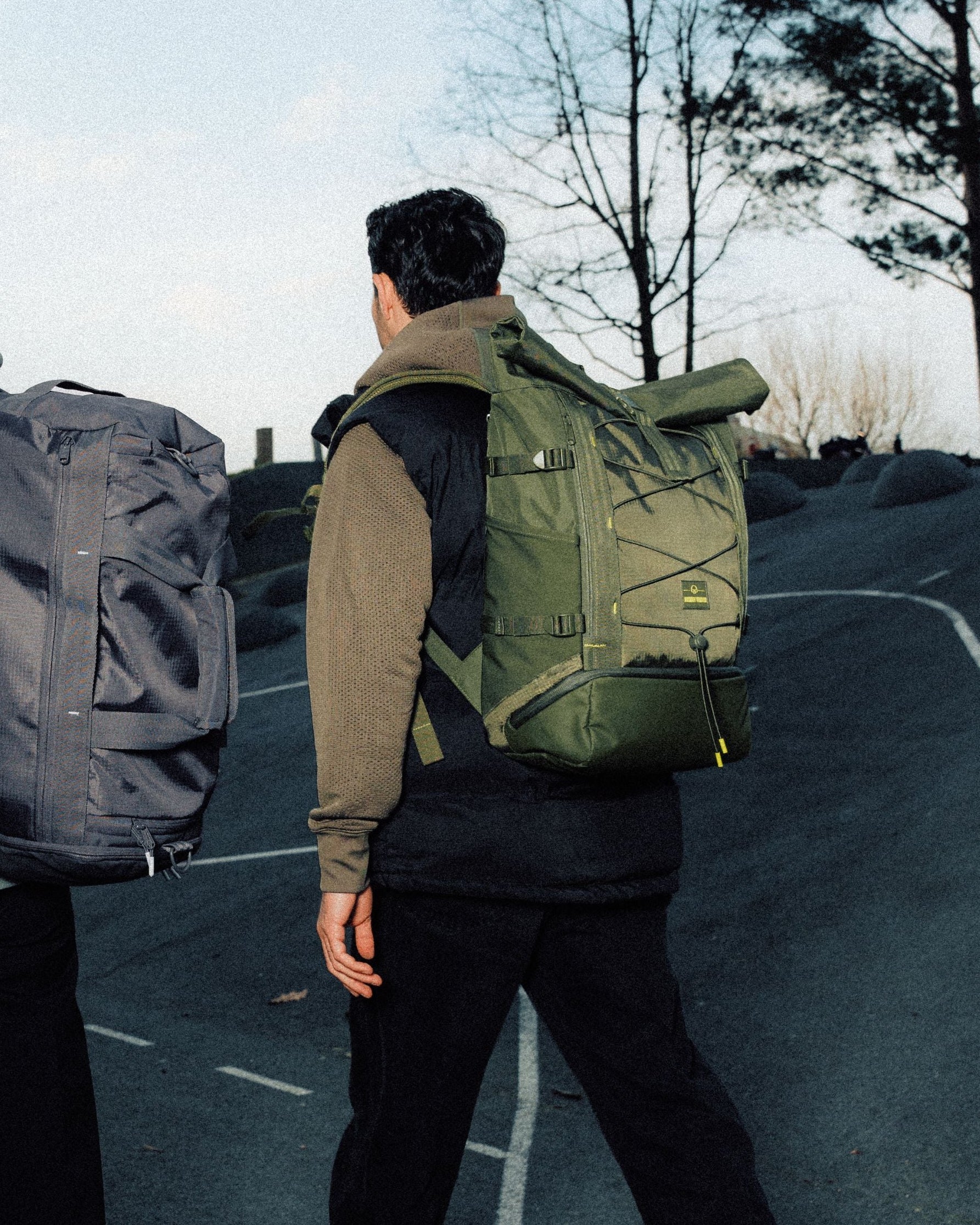 Zwei Personen gehen eine Straße entlang, tragen große Rolltop-Rucksäcke in Grau und Olivgrün