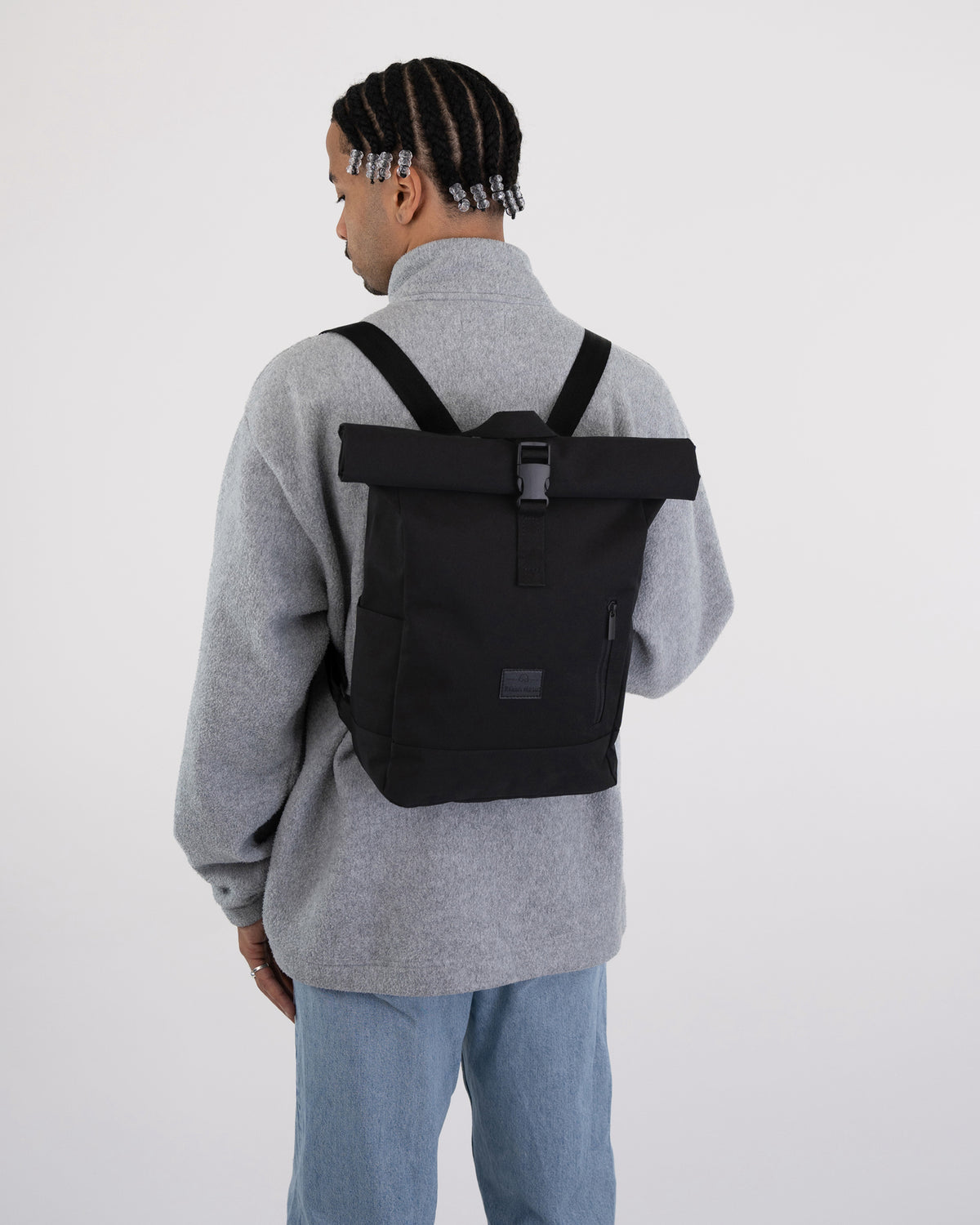 Schwarzer Rolltop Rucksack 'Robin Small' auf dem Rücken eines Mannes mit grauem Pullover und blauer Jeans