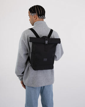 Schwarzer Rolltop Rucksack 'Robin Small' auf dem Rücken eines Mannes mit grauem Pullover und blauer Jeans