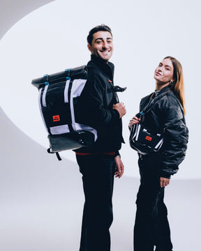 Mann mit schwarzem Rolltop Rucksack 'Ashton' und Frau mit passender Umhängetasche, beide in schwarzen Outfits