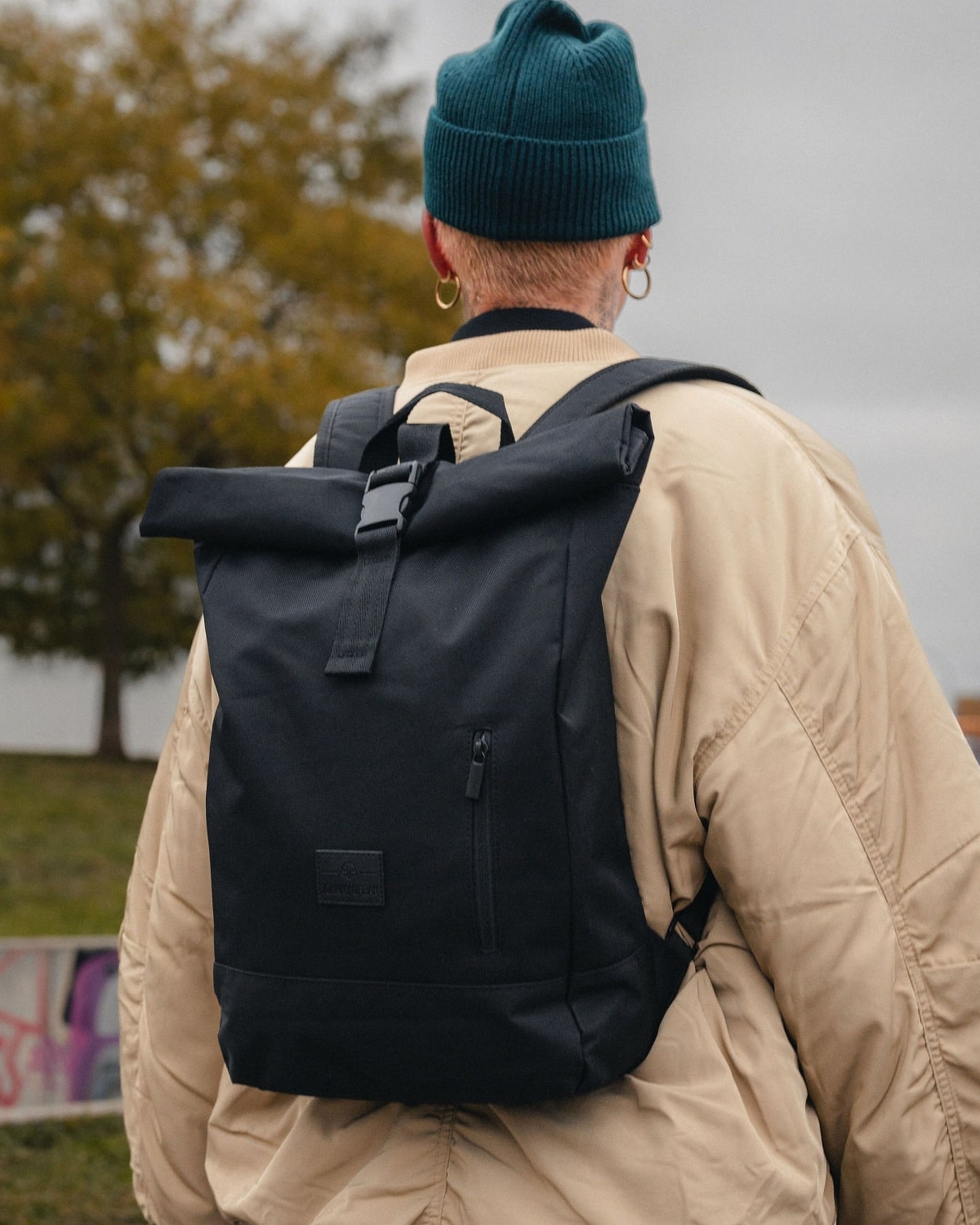 Dame trägt schwarzen Rolltop-Rucksack 'Robin Medium' von Johnny Urban mit Fronttasche, vor grauem Himmel und Herbstbaum