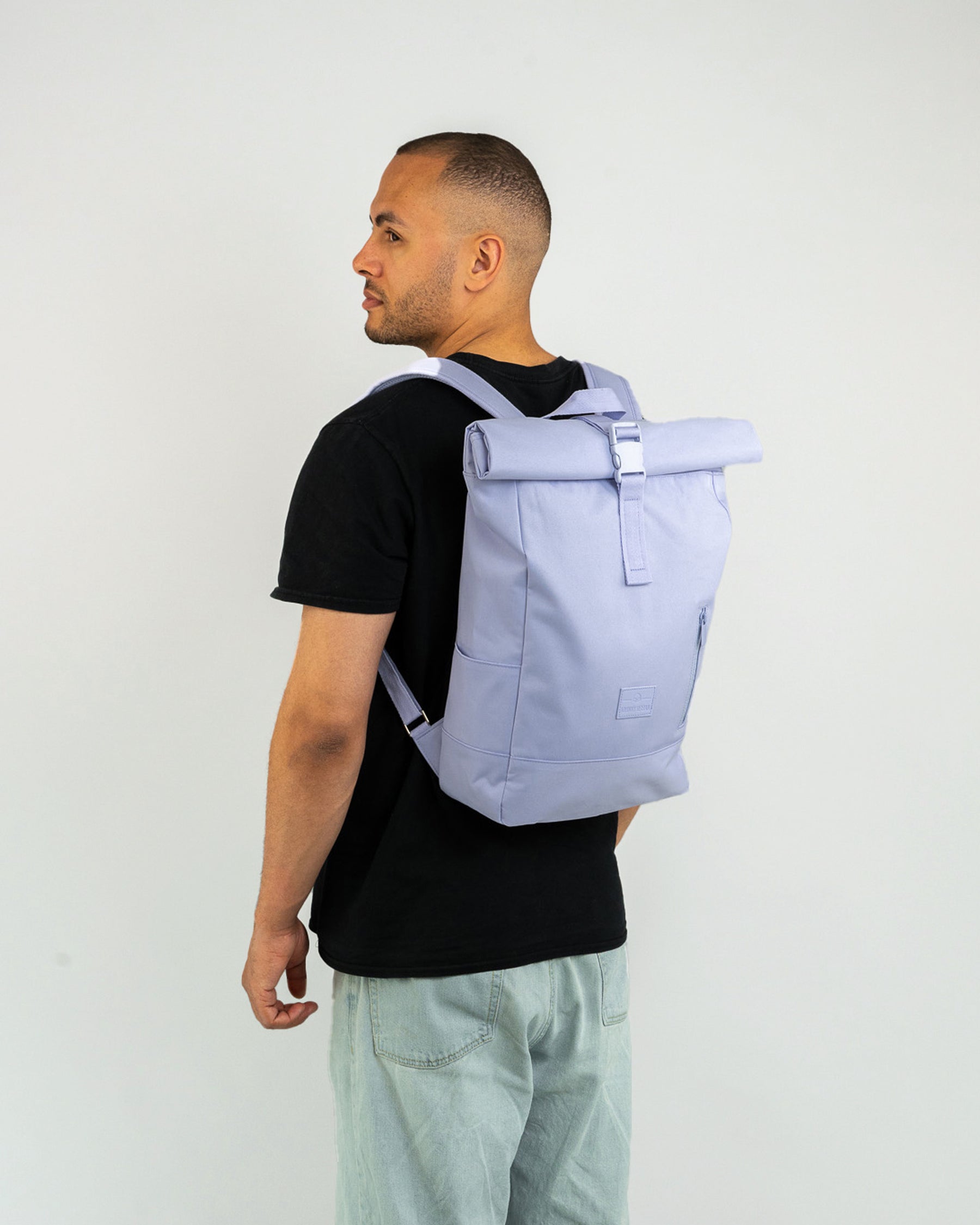 Mann trägt Johnny Urban Rolltop Rucksack 'Robin Medium' in flieder-lila mit schwarzem T-Shirt und hellen Hosen