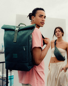 Mann mit grünem Johnny Urban Rolltop Rucksack 'Robin Large' auf dem Rücken und Frau mit grüner Umhängetasche urbanes Umfeld