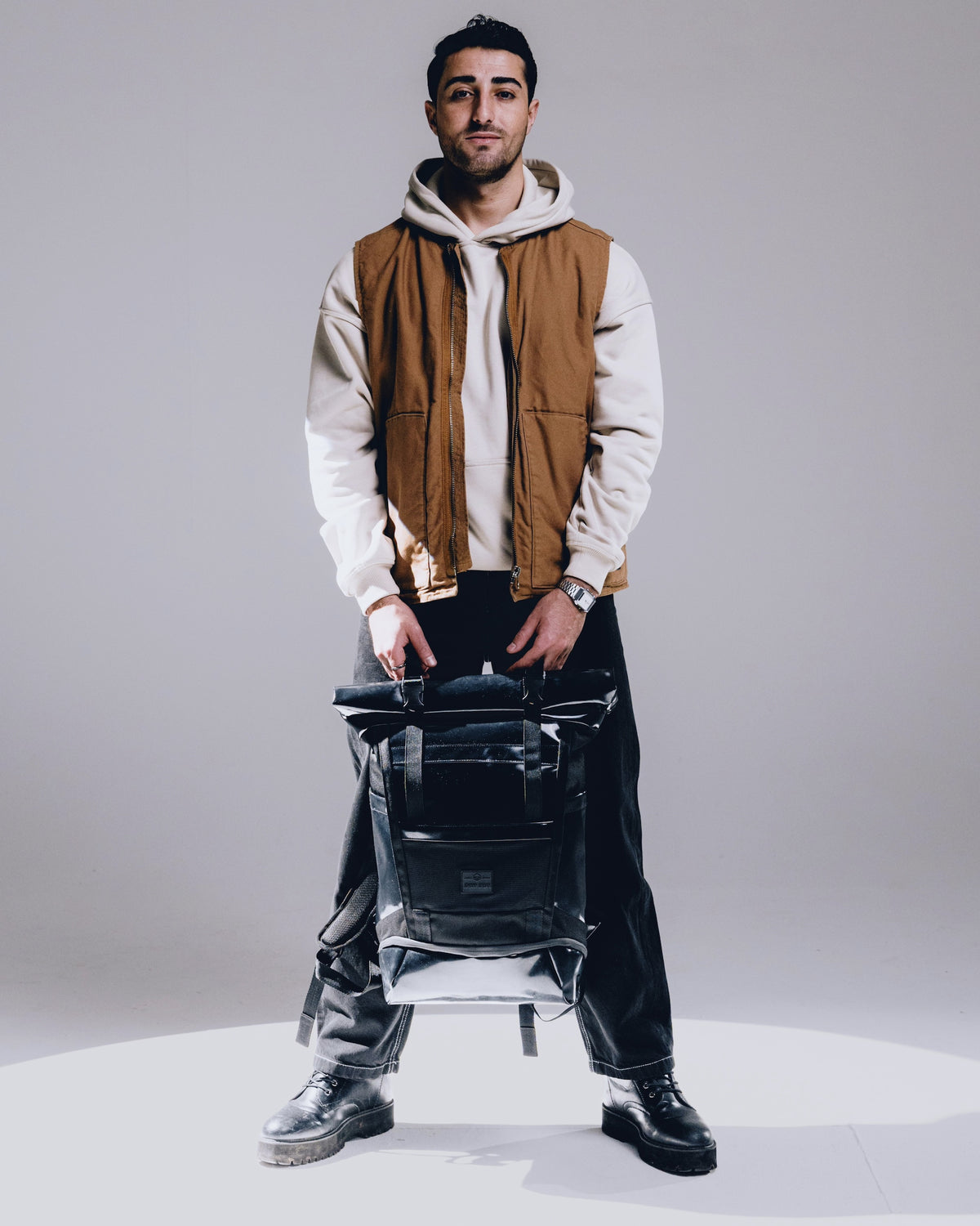 Mann mit beigem Kapuzenpullover und brauner Weste hält schwarzen Rolltop Rucksack 'Ashton' von Johnny Urban