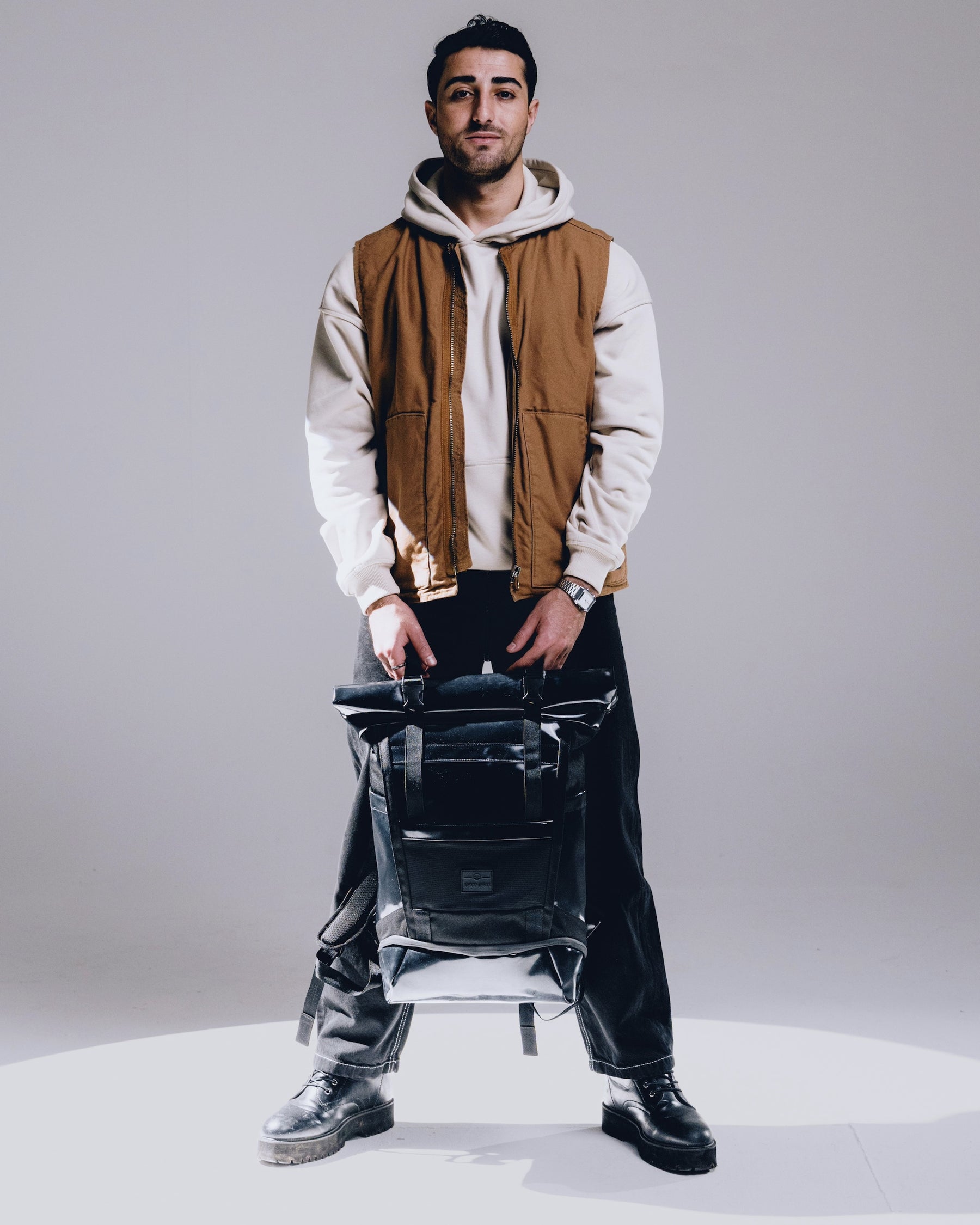 Mann mit beigem Kapuzenpullover und brauner Weste hält schwarzen Rolltop Rucksack 'Ashton' von Johnny Urban