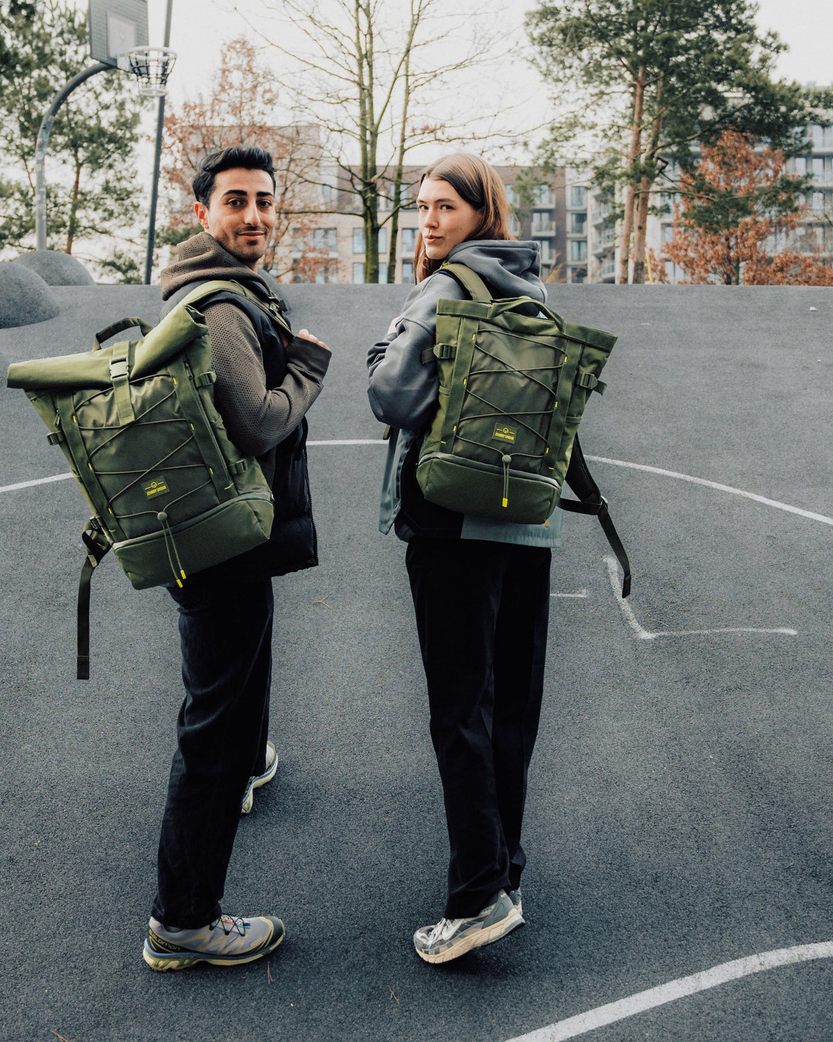 Zwei Personen, jeweils mit olivgrünem Rucksack 'Harper', auf einem urbanen Basketballplatz