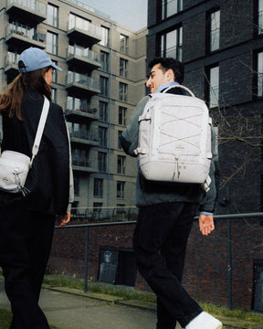 Zwei Personen gehen an einem urbanen Wohnhaus vorbei, eine trägt den beige Rucksack ‚Jack‘ von Johnny Urban