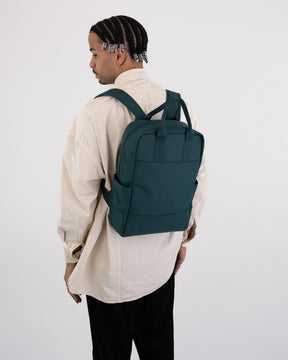 Mann mit Zöpfen trägt grünen Rucksack 'Hailey' aus Stoff, beige Hemd, schwarze Hose vor weißem Hintergrund