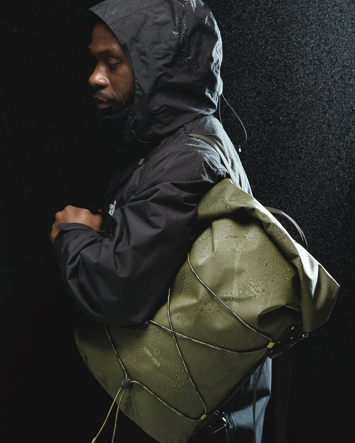 Mann in schwarzer Regenjacke trägt olivefarbenen rolltop Rucksack 'Myles' von Johnny Urban im Regen