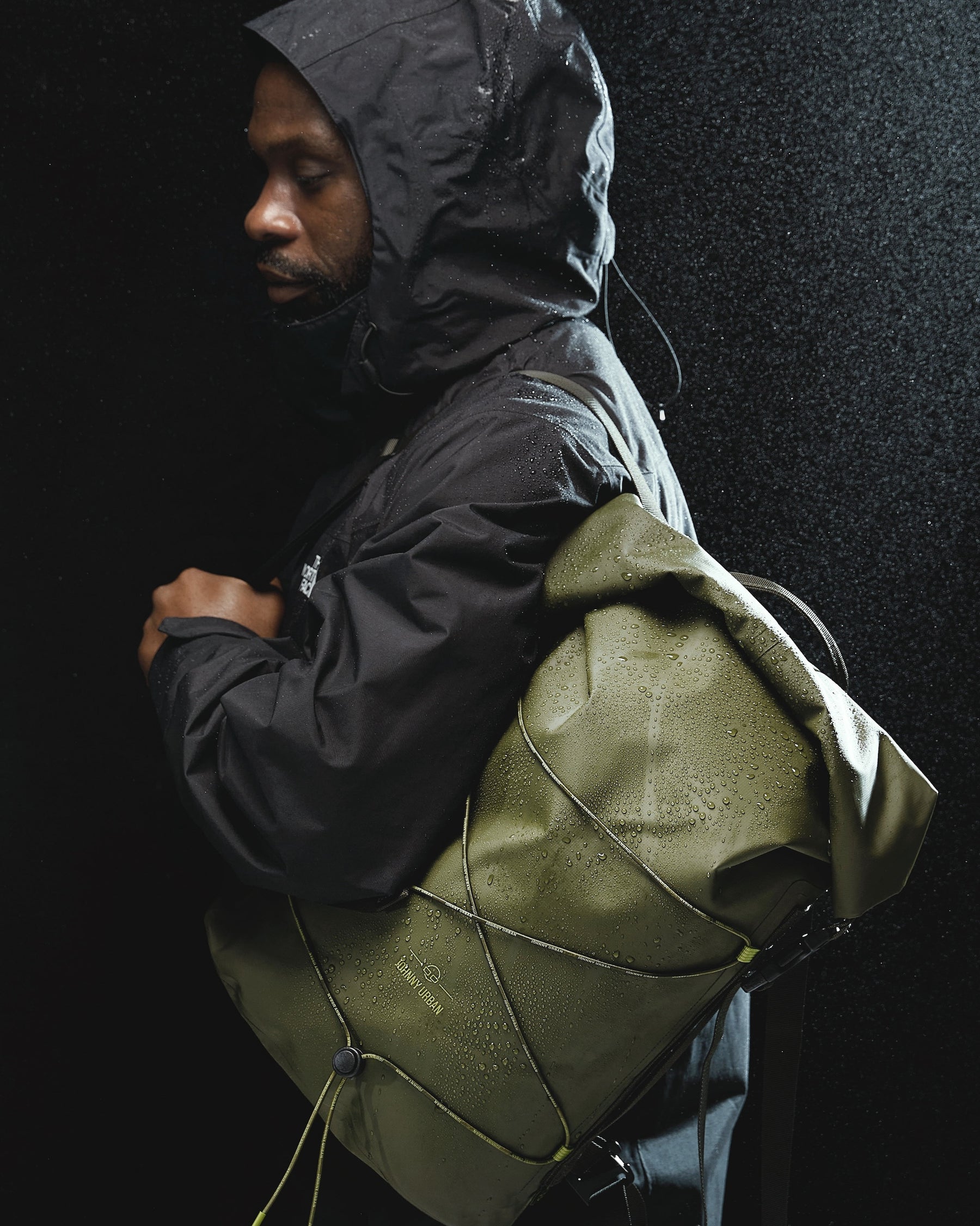 Mann in schwarzer Regenjacke trägt olivefarbenen rolltop Rucksack 'Myles' von Johnny Urban im Regen