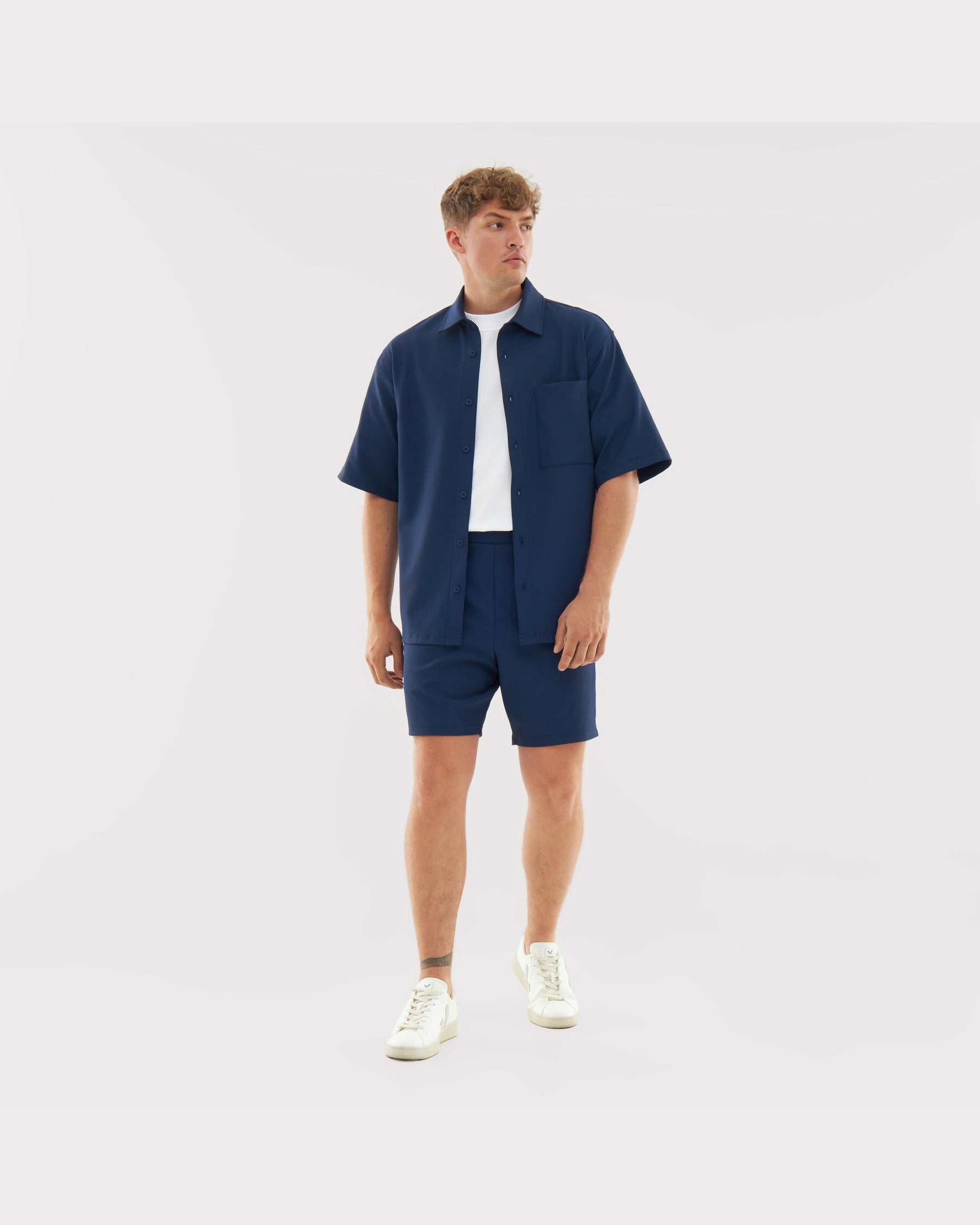 Männliches Modell trägt marineblaue Shorts 'Mason' und passendes Hemd, kombiniert mit weißem T-Shirt und weißen Sneakern
