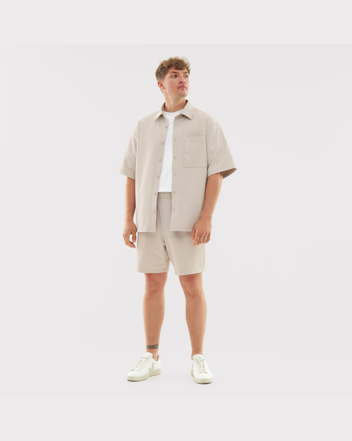 Männliches Modell trägt Sandfarbene Shorts und Kurzarm-Hemd aus dem Set 'Mason' mit weißen Turnschuhen