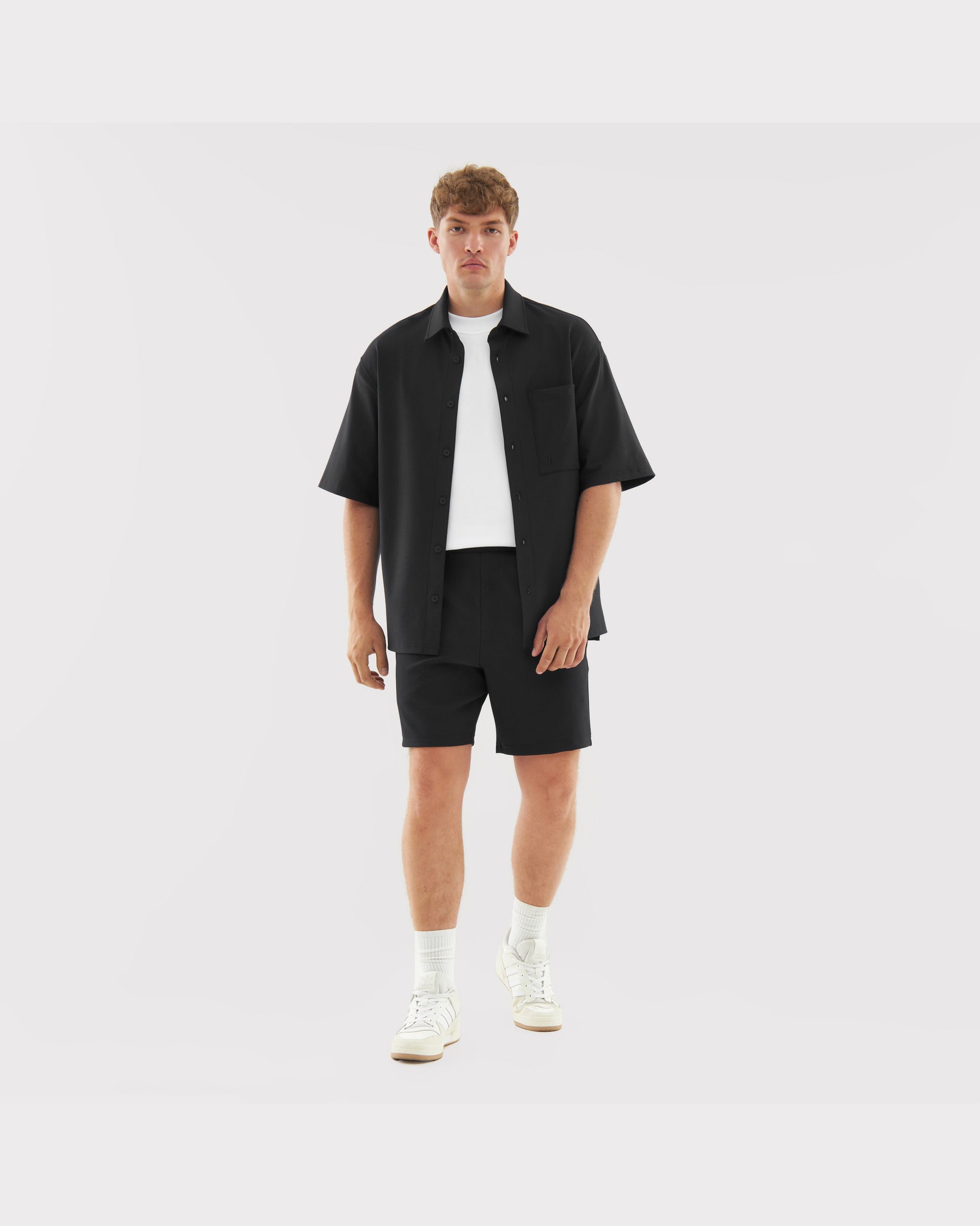 Männliches Modell trägt schwarze Shorts 'Mason', schwarze kurzärmelige Hemdjacke, weißes T-Shirt und weiße Sneakers