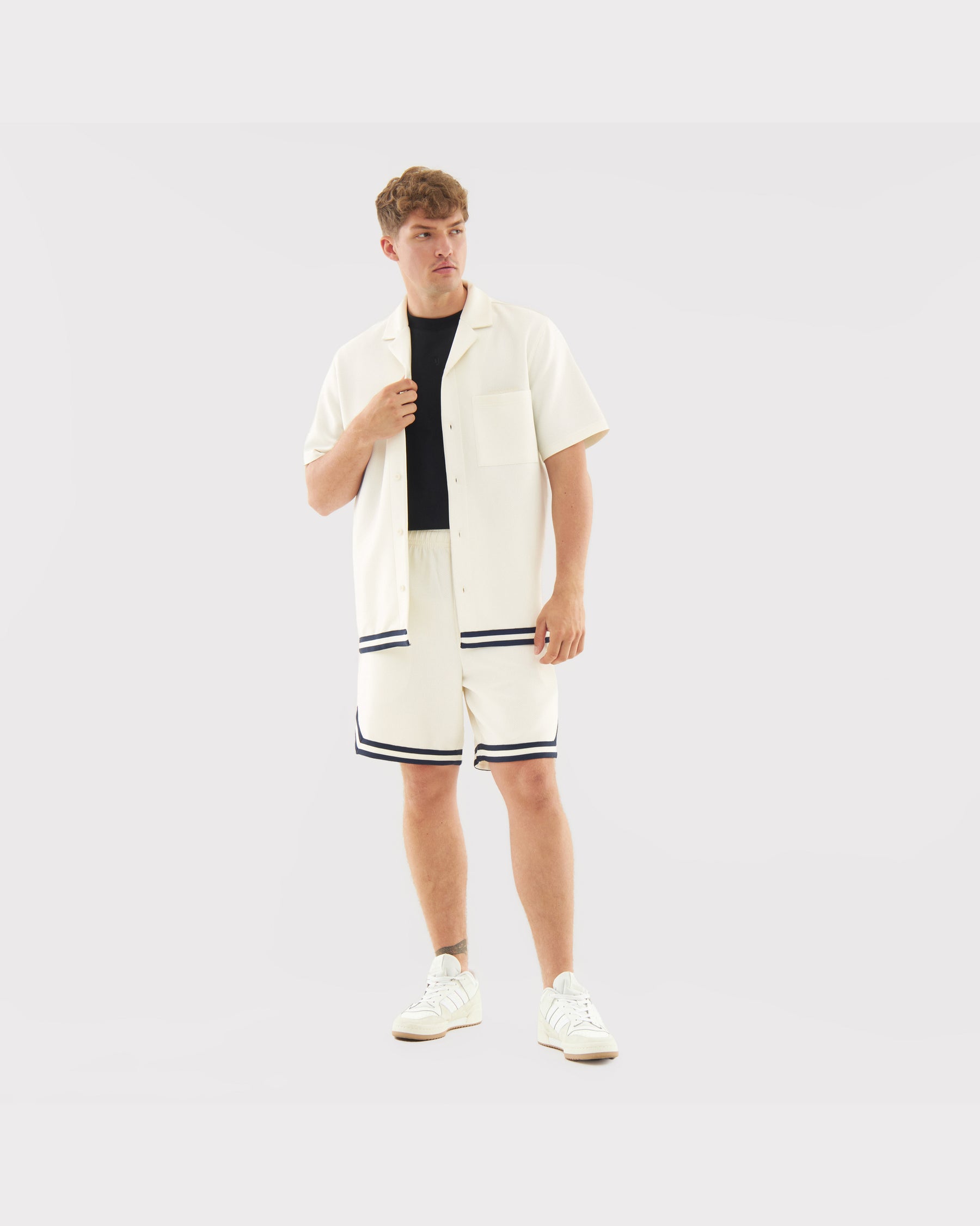Mann trägt cremefarbene Shorts 'Parker' mit schwarzem Saum kombiniert mit cremefarbener Kurzarmjacke und weißen Sneakern