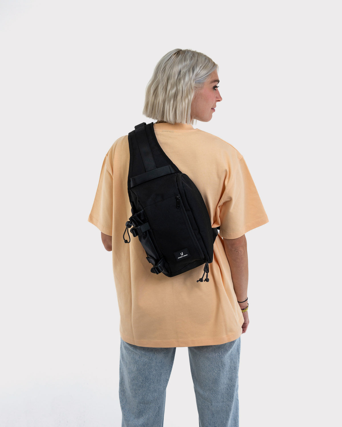 Frau mit kurzem blondem Haar trägt schwarze Johnny Urban Sling Bag 'Charlie' über beigem T-Shirt und blauer Jeans