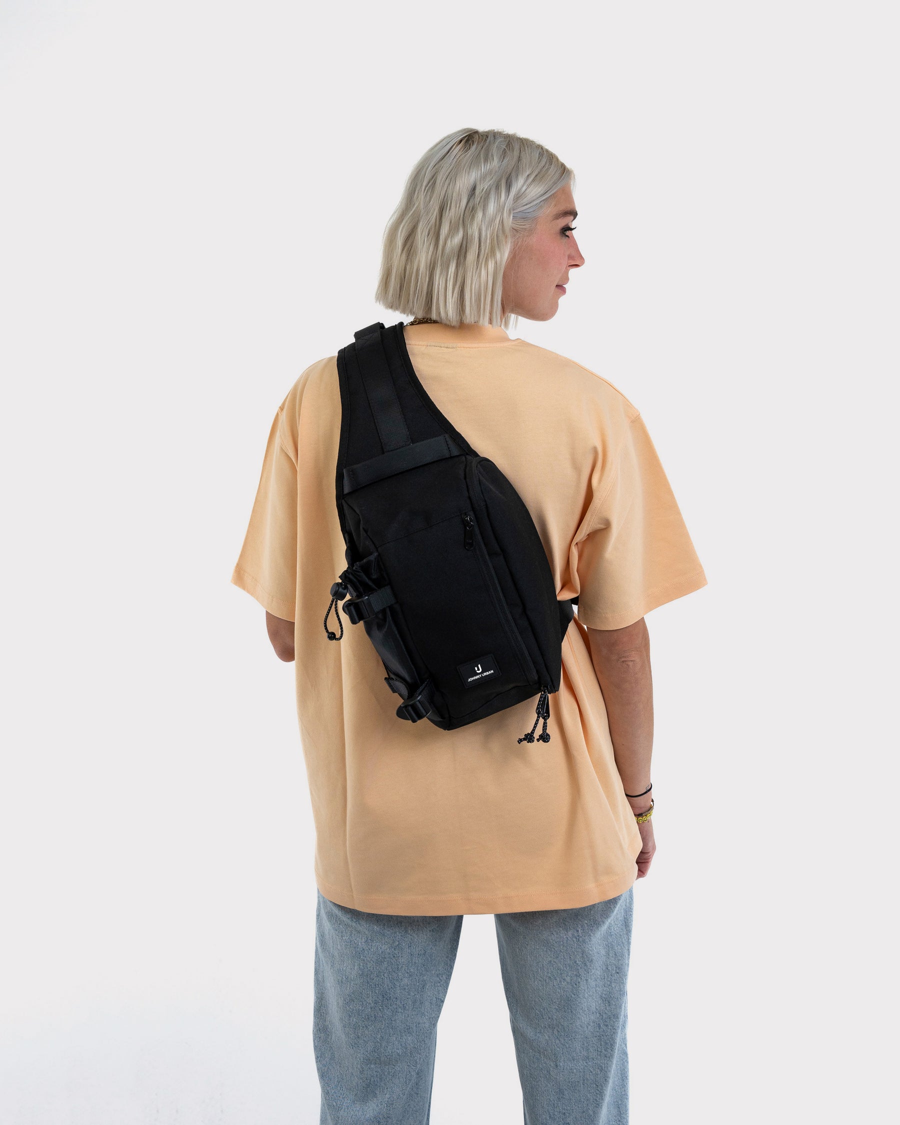 Frau mit kurzem blondem Haar trägt schwarze Johnny Urban Sling Bag 'Charlie' über beigem T-Shirt und blauer Jeans