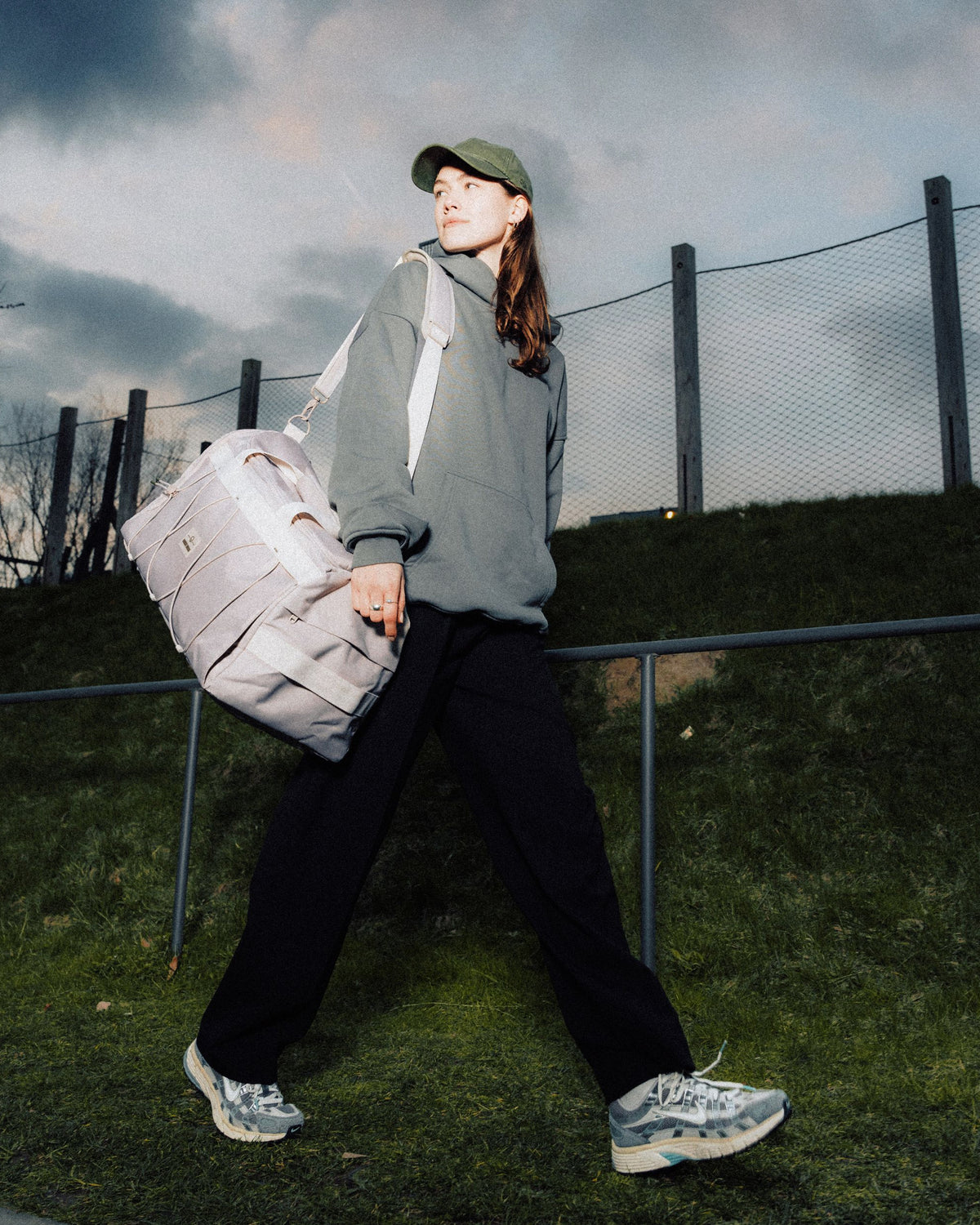Frau trägt beige Duffle Bag "Shawn Medium" über der Schulter, trägt grünen Pullover, schwarze Hose und Baseballcap