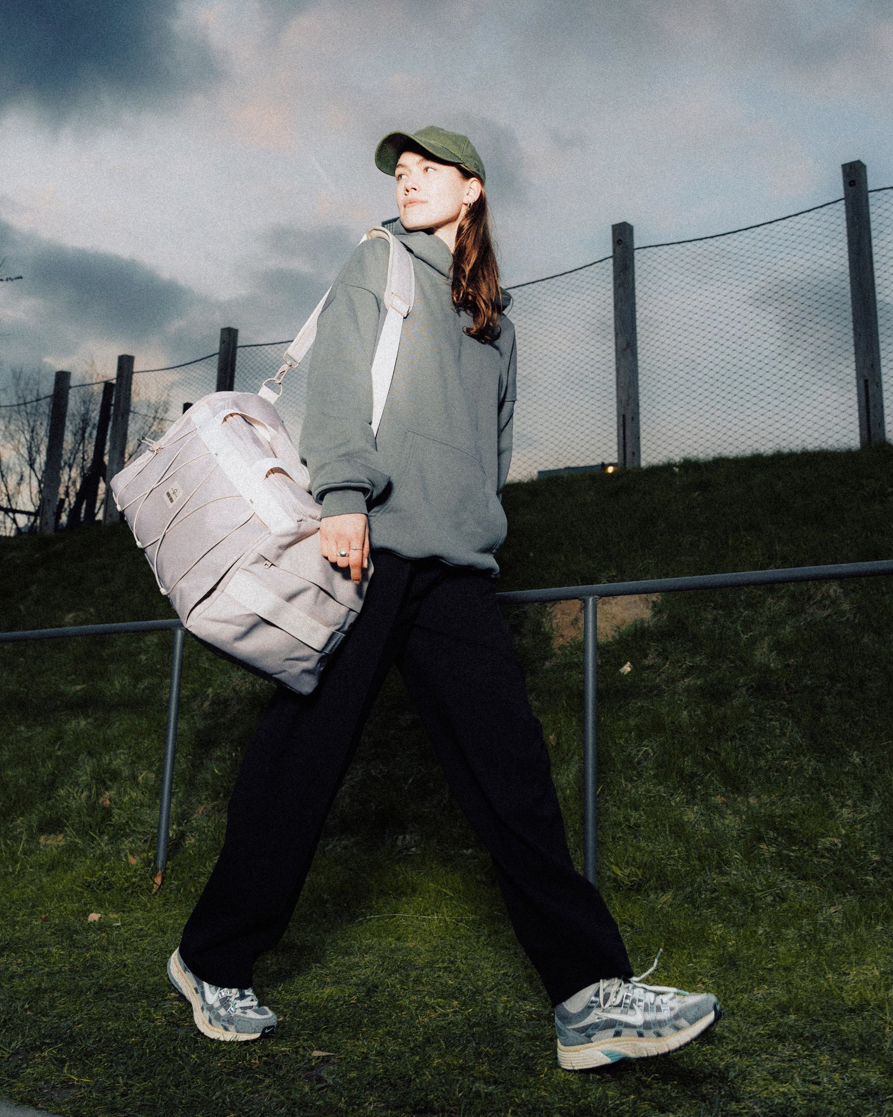 Frau trägt beige Duffle Bag "Shawn Medium" über der Schulter, trägt grünen Pullover, schwarze Hose und Baseballcap