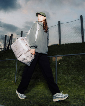Frau trägt beige Duffle Bag "Shawn Medium" über der Schulter, trägt grünen Pullover, schwarze Hose und Baseballcap