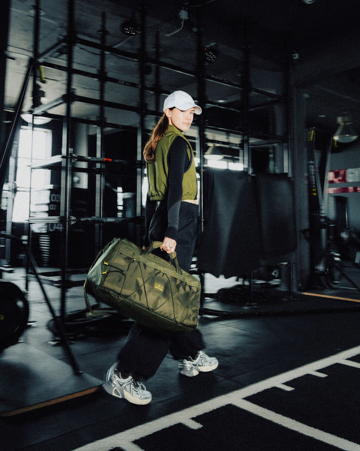 Frau in schwarzer Kleidung und olivfarbener Weste trägt Duffle Bag 'Shawn Medium' in Olivgrün im Fitnessstudio