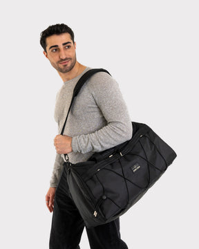 Mann trägt schwarze Johnny Urban Sporttasche 'Shawn Medium' über Schulter, trägt grauen Pullover und schwarze Hose