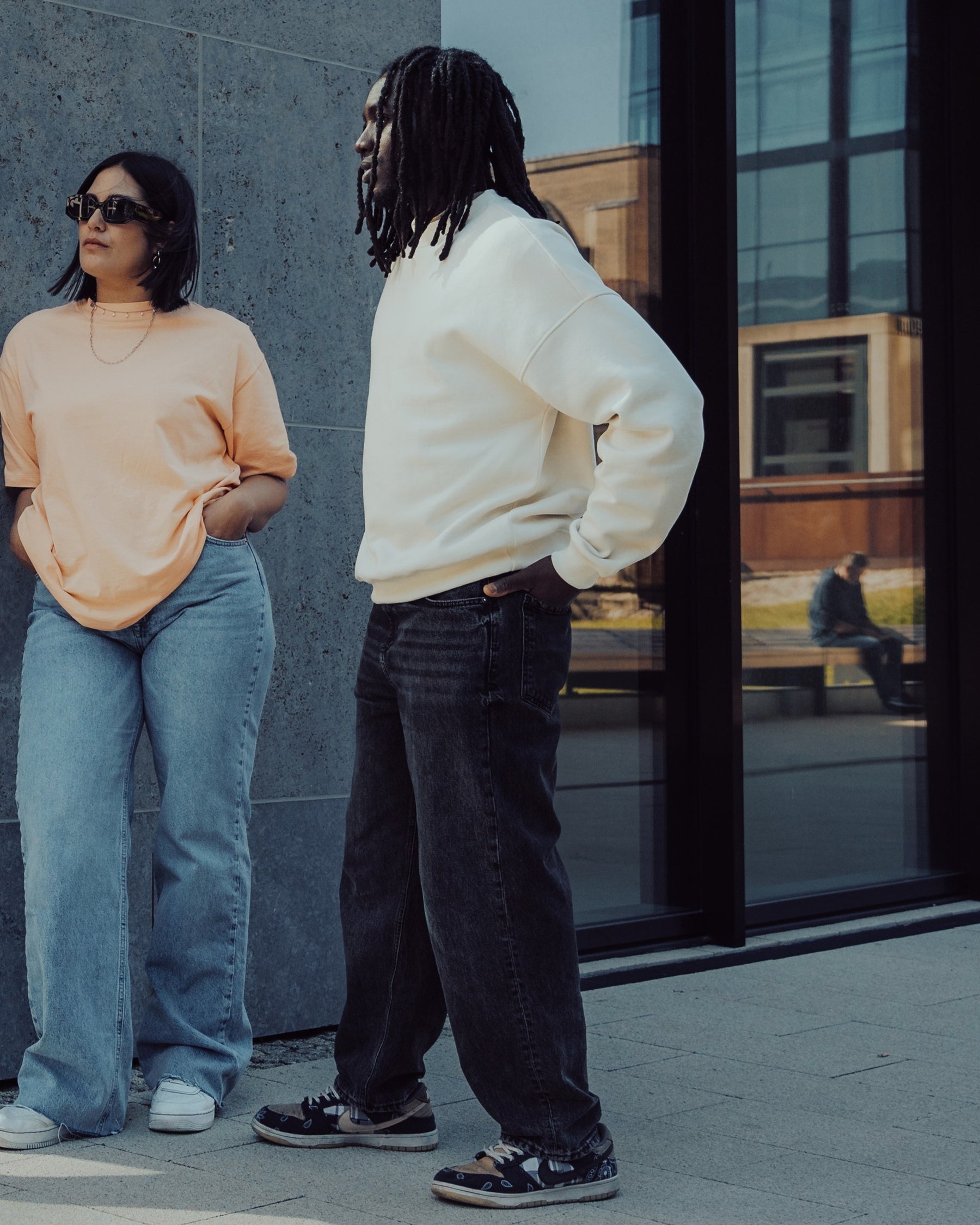 Zwei Personen vor modernem Gebäude, eine trägt weißen Oversized Crewneck Pullover 'Carter', die andere ein beiges T-Shirt
