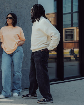 Zwei Personen vor modernem Gebäude, eine trägt weißen Oversized Crewneck Pullover 'Carter', die andere ein beiges T-Shirt