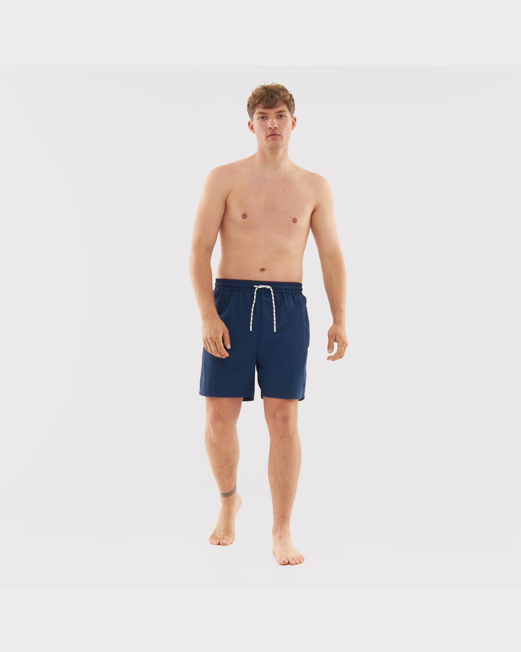 Mann trägt navyblaue Badehose 'Jay' mit weißem Kordelzug, barfuß vor weißem Hintergrund