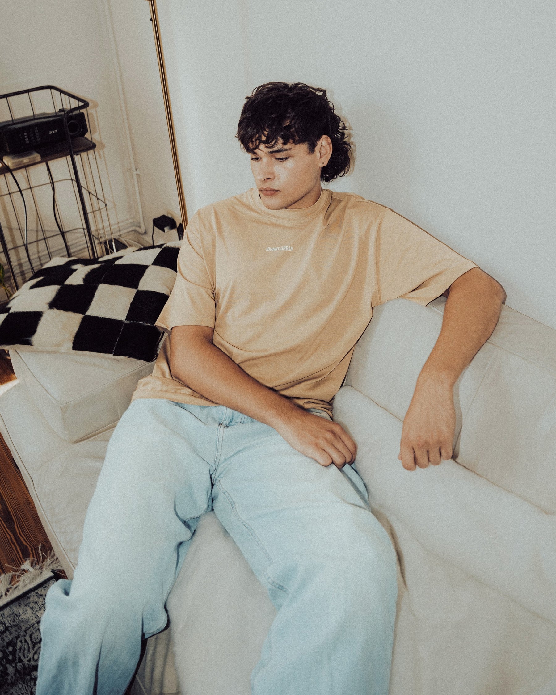 Mann sitzt auf weißem Sofa trägt oversized beige T-Shirt 'Sammy Johnny Urban' und helle Jeans