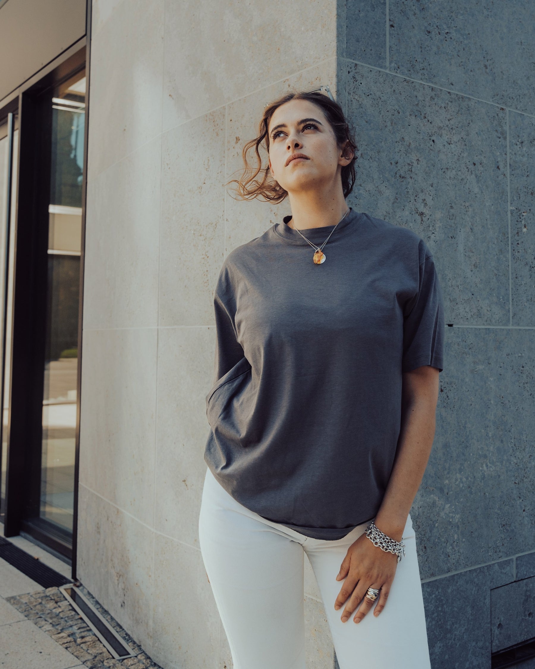Frau trägt das graue Oversize T-Shirt 'Sammy' von Johnny Urban und weiße Hosen, steht vor einer grauen Steinwand