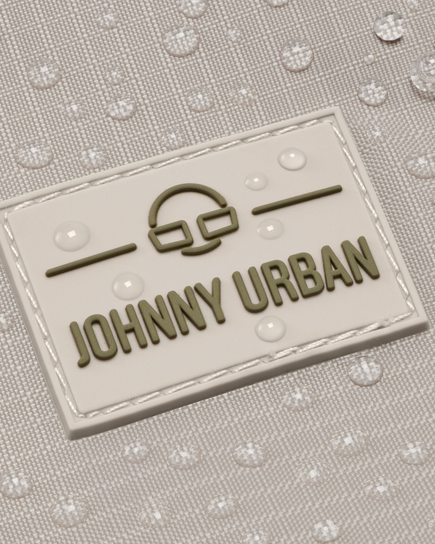 Detailansicht des Johnny Urban Logos auf einem wasserabweisenden, beigefarbenen Rucksack Harper mit Wassertropfen
