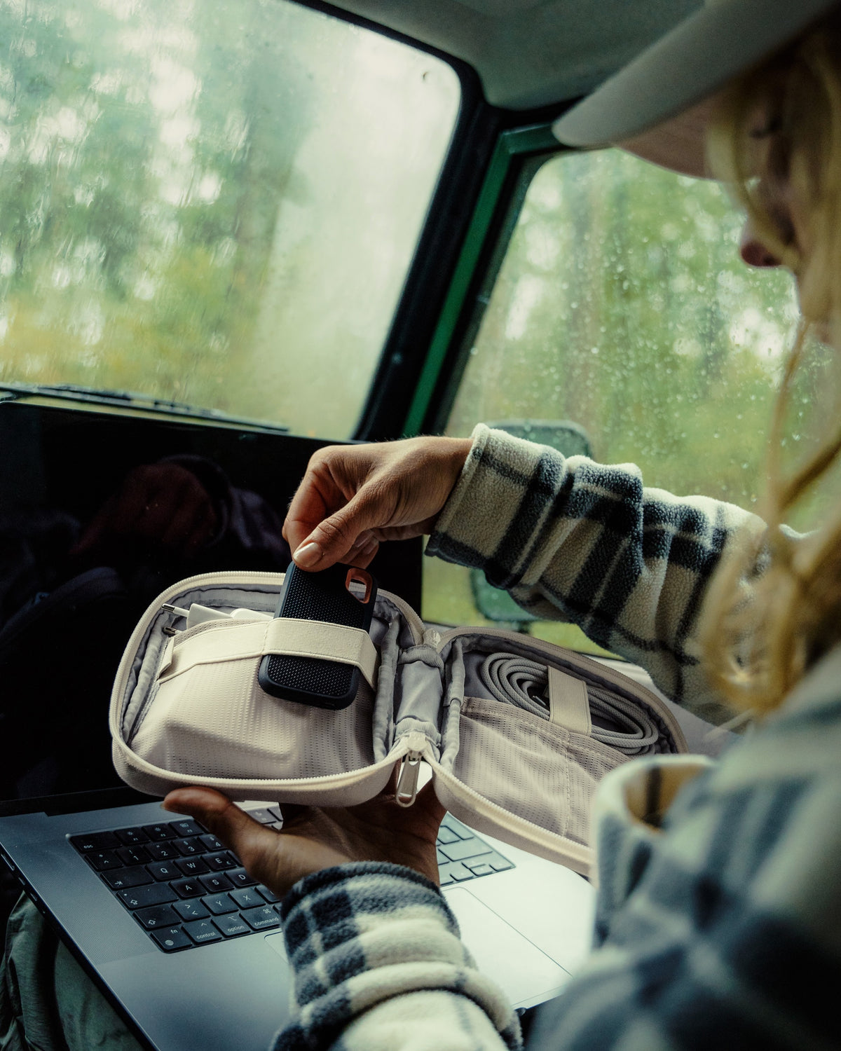 Person in kariertem Hemd nutzt den Tech Organizer 'Ted' zum Verstauen eines Geräts neben einem Laptop im Auto