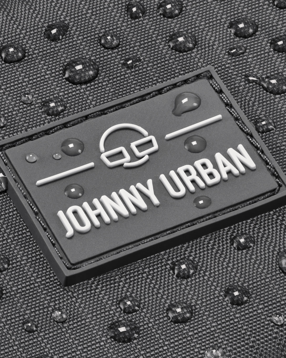 Nahaufnahme des Johnny Urban Logos auf grauem, wasserabweisendem Stoff mit Wassertropfen