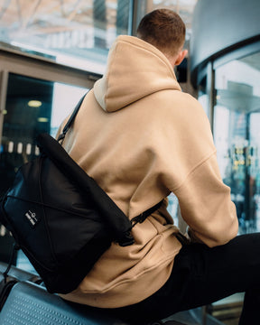Schwarze Johnny Urban Umhängetasche 'Dylan' aus robustem Material, getragen von einem Mann im beigen Hoodie in moderner Umgebung