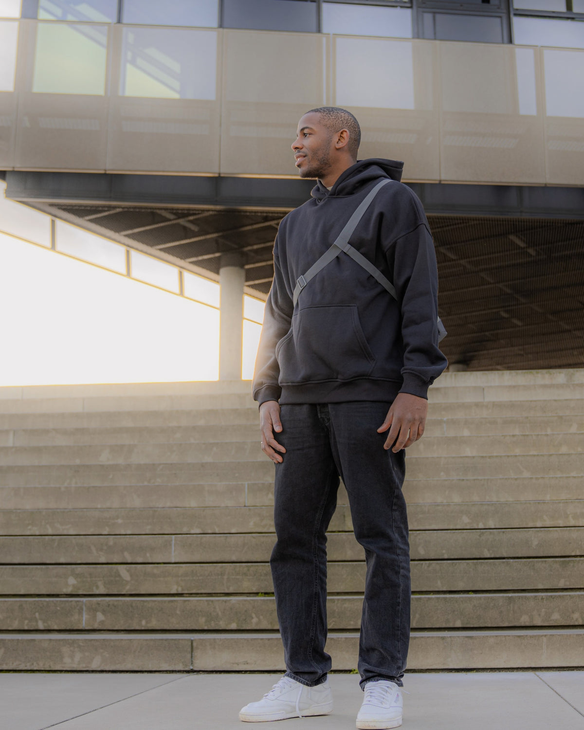Mann trägt schwarze Umhängetasche 'Dylan' von Johnny Urban, schwarzen Hoodie, Jeans und weiße Sneaker vor modernen Treppen